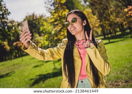 Photo of attractive asian lady fingers make v-sign take selfie beaming toothy smile wear yellow leather coat day outdoors