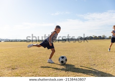 Biracial elementary schoolboy kicking soccer ball while playing on field against sky. unaltered, childhood, education, sports training, team sport and sports activity concept. Royalty-Free Stock Photo #2138463807