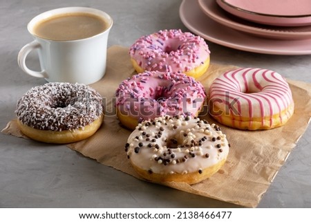 Several donuts lie on craft parchment paper on gray surface. Porcelain cup of coffee and pink plates stands next to donuts laid out on table. Selective focus.