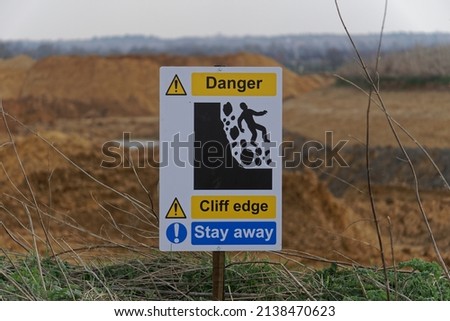 A yellow and blue warning sign with an image of a person falling and the words Danger Cliff Edge Stay Away on the perimeter of quarry workings in the English countryside