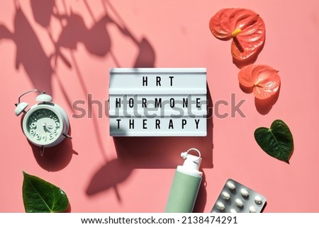 Text HRT Replacement Therapy on light box. Menopause, hormone therapy concept. Oestrogen replacement therapy awareness. Pink background with alarm clock, exotic leaves, pills, estrogene gel. Royalty-Free Stock Photo #2138474291