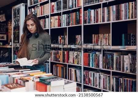Young female customer reading a book in bookstore while buying some good literature. Woman picking a book to read. Royalty-Free Stock Photo #2138500567