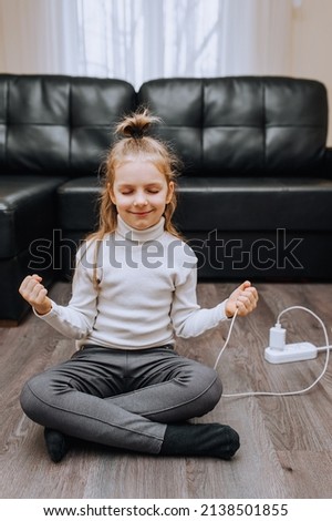 A beautiful, little red-haired girl, a child, a smiling robot sits on the floor and is charged from a charger with energy through electricity from a white cord. Photography, concept.
