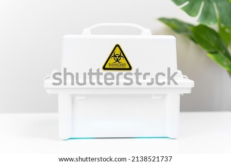 White plastic box with handle and sticker warning of threat of biological contamination is on the table in medical lab, front view. Container for the disposal of biowaste