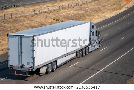 Industrial bonnet gray big rig semi truck with aerodynamic roof spoiler transporting commercial cargo in dry van semi trailer running on the divided summer highway road with yellow grass on the hill Royalty-Free Stock Photo #2138536419