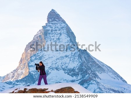 Interested woman photographer standing on snow covered rock with Matterhorn peak in background capturing scenic mountain views of Swiss Alps on winter day