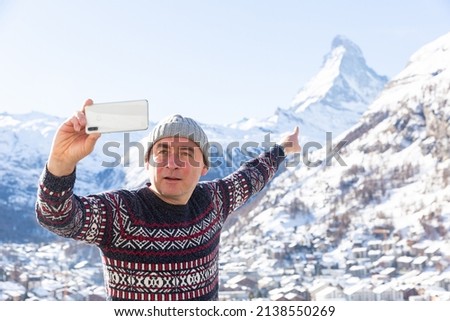 Positive travel blogger using smartphone to record vlog while hiking in Swiss Alps in wintertime, pointing at snow-capped Matterhorn peak in background