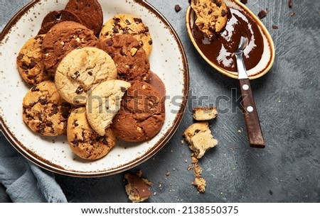 Assorted chocolate chip cookies on wooden plate with cup of coffee on dark background, top view.