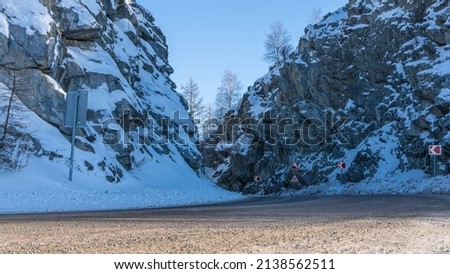 The road meanders between steep cliffs. Snow on the rocks. Warning road signs along the bend of the highway. Altai. 