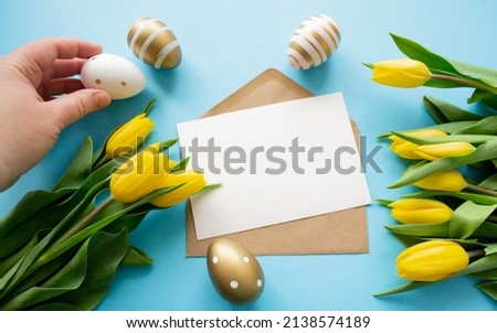 
Easter: colored eggs, yellow tulips. Letter  with copy space, blue background. Hand holds white Easter egg Royalty-Free Stock Photo #2138574189