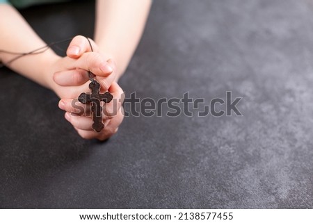 Child's hands clasped in prayer and holding the wooden cross on dark background.