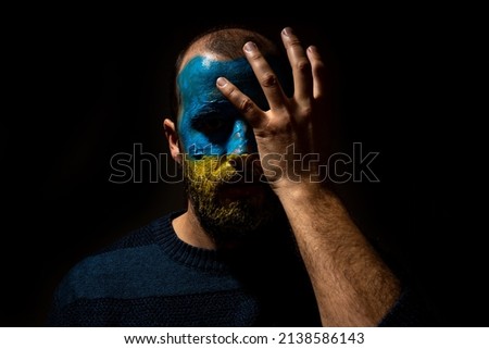 Stop war, conflict between Ukraine and Russia. Portrait of a young man with his face painted in the blue and yellow colors of the flag. Very sad with a hand on his face