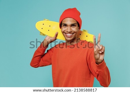 Young smiling happy african american man 20s in orange shirt hat hold skateboard yellow penny board show v-sign isolated on plain pastel light blue background studio portrait. People lifestyle concept