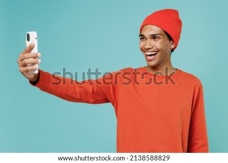 Young happy african american man in orange shirt hat doing selfie shot on mobile cell phone post photo on social network isolated on plain pastel light blue background studio. People lifestyle concept