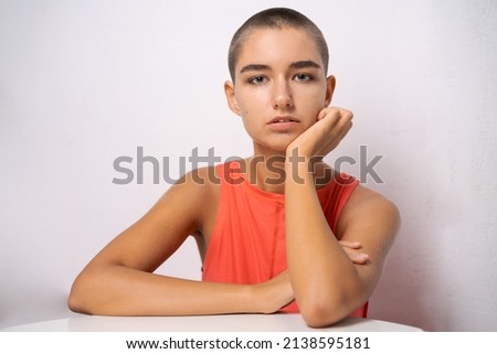 Caucasian girl with short hair, almost bald, holds her hands behind her head, leaning on the table in alarm in an orange T-shirt on a light background close-up. The concept of illness and loneliness