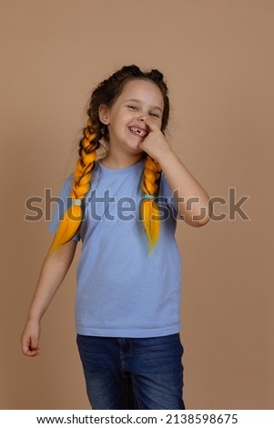 Caucasian naughty small girl smiling shining picking nose with finger looking at camera having kanekalon braids on beige background wearing blue t-shirt. Bad habits.