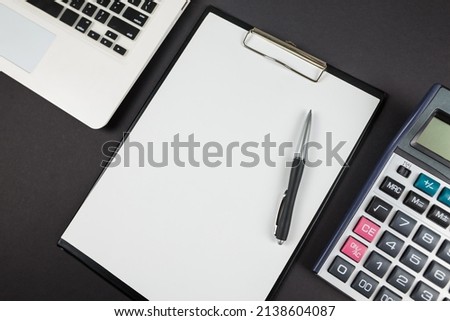 Top view photo of black clipboard folder with white paper sheet, pen, calculator and laptop on isolated black background.