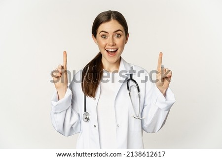 Enthusiastic young woman doctor smiling, pointing fingers up, wearing hospital uniform, standing over white background