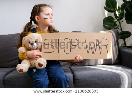 Portrait of upset little girl child holding banner with inscription Stop WAR, sitting alone on grey sofa at home with fluffy bear. Kid with flag of Ukraine on face. Ukrainian geopolitics globe crisis