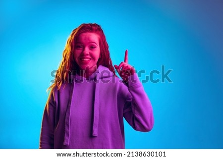 Portrait of young stylish girl rising finger expressing idea isolated over blue studio background in pink neon light. Concept of emotions, facial expression, youth, aspiration. Copy space for ad