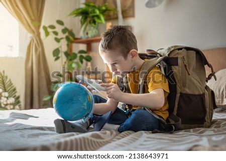 A child boy with a large backpack is playing with a toy airplane and a globe of the earth. The concept of going on a journey towards adventure.