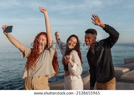 Taking happy selfies by the sea. Group of three friends cheering and dancing in front of a camera phone. Multicultural friends having a good time while hanging out together next to the sea.