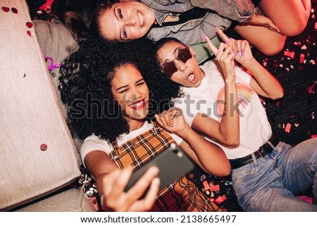 Selfies on girls night out. High angle view of three happy friends taking a selfie while lying on the floor at a house party. Group of cheerful female friends having fun together on the weekend.