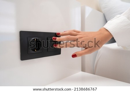 Close-up of a woman's hand with red nail polish turns on the light near the bed with a black switch on a white wall, next to the outlet Royalty-Free Stock Photo #2138686767