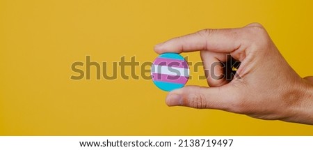 a young caucasian person holding a pin button patterned with the transgender pride flag, on a yellow background, in a panoramic format to use as web banner Royalty-Free Stock Photo #2138719497