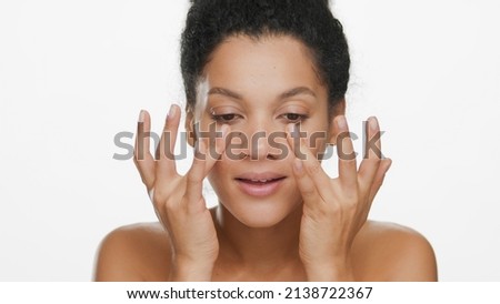 Close-up beauty portrait of young African American woman with bare shoulders touches her face skin under her eyes on white background | Droopy eyes removal concept Royalty-Free Stock Photo #2138722367
