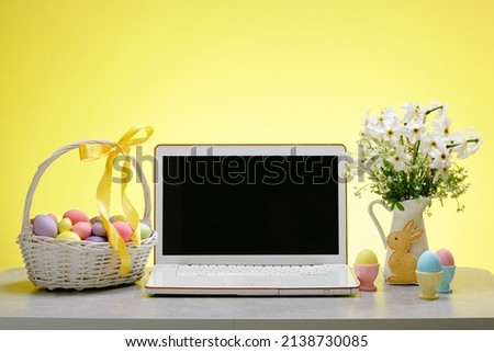 A laptop with a blank screen on a table festively decorated for the Easter holiday. Adorned working place with flowers and a basket with colorful eggs Royalty-Free Stock Photo #2138730085
