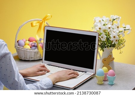 A womans hands on laptop with a blank screen on a table festively decorated for the Easter holiday. Adorned working place with flowers and a basket, with colorful eggs Royalty-Free Stock Photo #2138730087