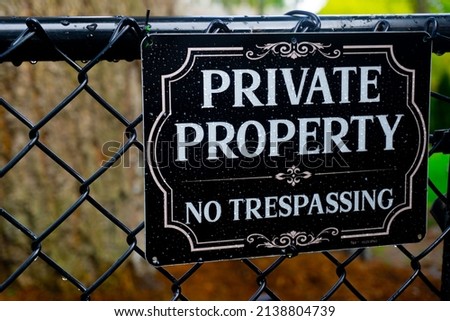 A sign reading Private Property No Tresspassing sits on a chain link fence in front of a natural background of trees and grass.