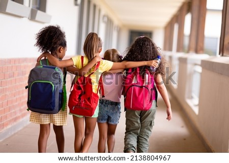 Rear view of multiracial elementary schoolgirls with backpacks and arm around walking in corridor. unaltered, childhood, together, education and back to school concept. Royalty-Free Stock Photo #2138804967