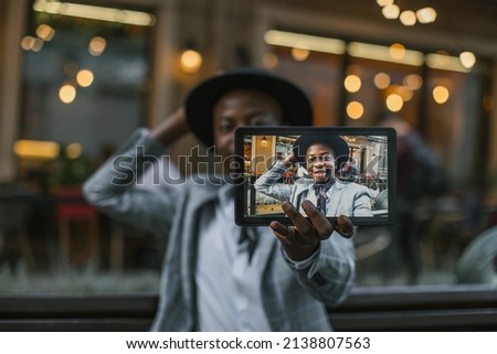 Close up of digital tablet with happy stylish african man on screen. Handsome young guy in hat and suit taking selfie while sitting on bench.