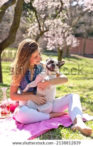 Panoramic view of woman tickling dog on picnic blanket at park in springtime. Vertical full view of young woman being playful with french bulldog at park in summer. Animals and people lifestyle.