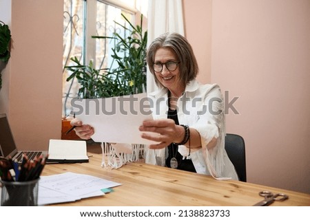 Inspired elderly business woman female fashion designer looks at drawn sketches for clothes while sitting at wooden table in her design studio office or workshop and smiling with cheerful toothy smile