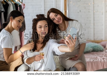Group of three female friends posing together taking photo use smartphone at home party. Happy women making selfie on mobile phone having fun drinking champagne at home bedroom interior