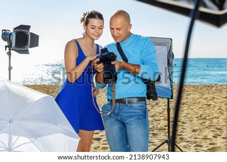 Photographer and positive smiling model discussing picture on camera display during photo session on seaside in sunny