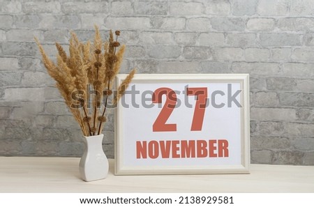 november 27. 27th day of month, calendar date. White vase with ikebana and photo frame with numbers on desktop, opposite brick wall. Concept of day of year, time planner, autumn month