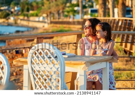 Mother daughter meet the morning sunrise sitting at a table outdoors by the sea. Summer holiday, rest, travel concept.