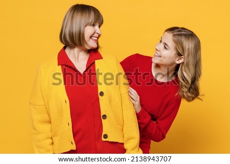Happy smiling cool woman 50s wearing red shirt have fun with teenager girl 12-13 years old. Grandmother granddaughter look to each other isolated on plain yellow background. Family lifestyle concept