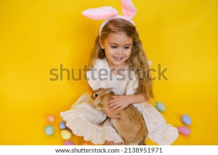 cute smiling blonde woman with bunny ears holding a red rabbit in her hands, on a yellow studio background, space for text, a child celebrates easter. High quality photo