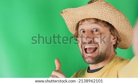 Close up young smiling happy caucasian man yellow sweatshirt farmer hat doing selfie shot showing thumb up sign isolated on plain green background studio portrait. People lifestyle concept