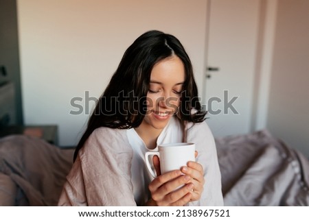Lovely adorable girl with dark hair is holding a cup of coffee and covering by blanket in the bedroom in morning in sunshine. Home concept. Morning wake up at home. High quality photo