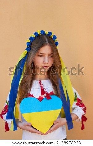 
A little Ukrainian girl in a traditional costume holds a heart with the flag of Ukraine. Pray for Ukraine. Save Ukraine. The children are asking for peace. There is no war.