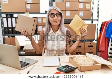 Middle age grey-haired woman holding package and envelope at warehouse smiling looking to the side and staring away thinking. 
