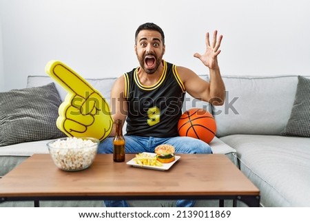 Young hispanic man with beard holding basketball ball cheering tv game celebrating victory with happy smile and winner expression with raised hands 