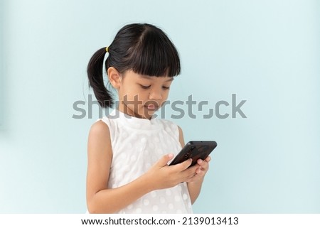 Asian kid girl A cute face with black hair,6 years old,wearing a white shirt,standing in a room with a light blue background.and is bent over to watch her favorite animation using a black smartphone.