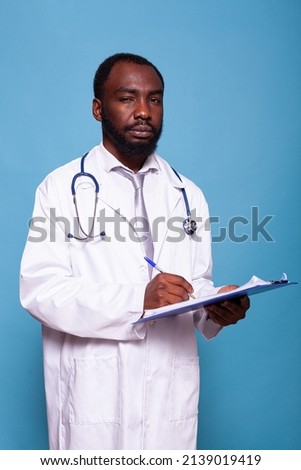 Portrait of overconfident doctor looking at camera holding clipboard with patient charts. Confident medic posing in lab coat with stethoscope and writing on medical history papers.
